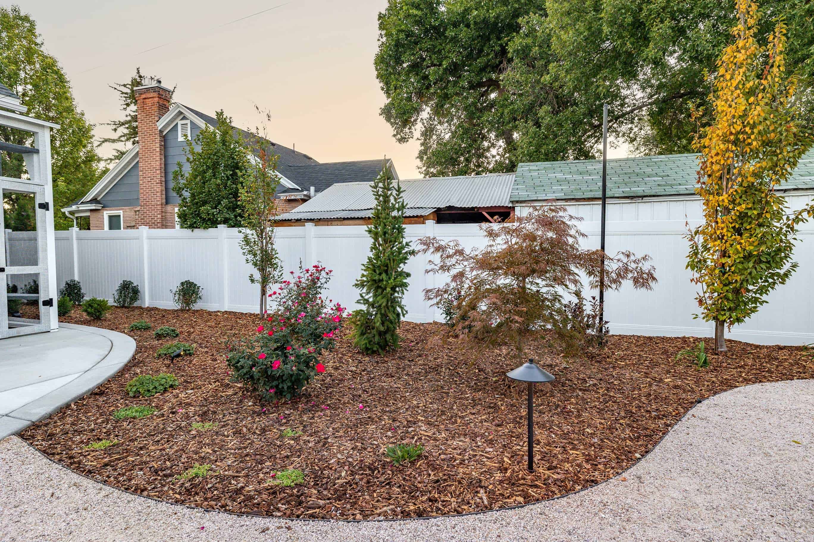 Mulched planting bed with flowering shrubs, young trees, gravel path, and landscape lighting