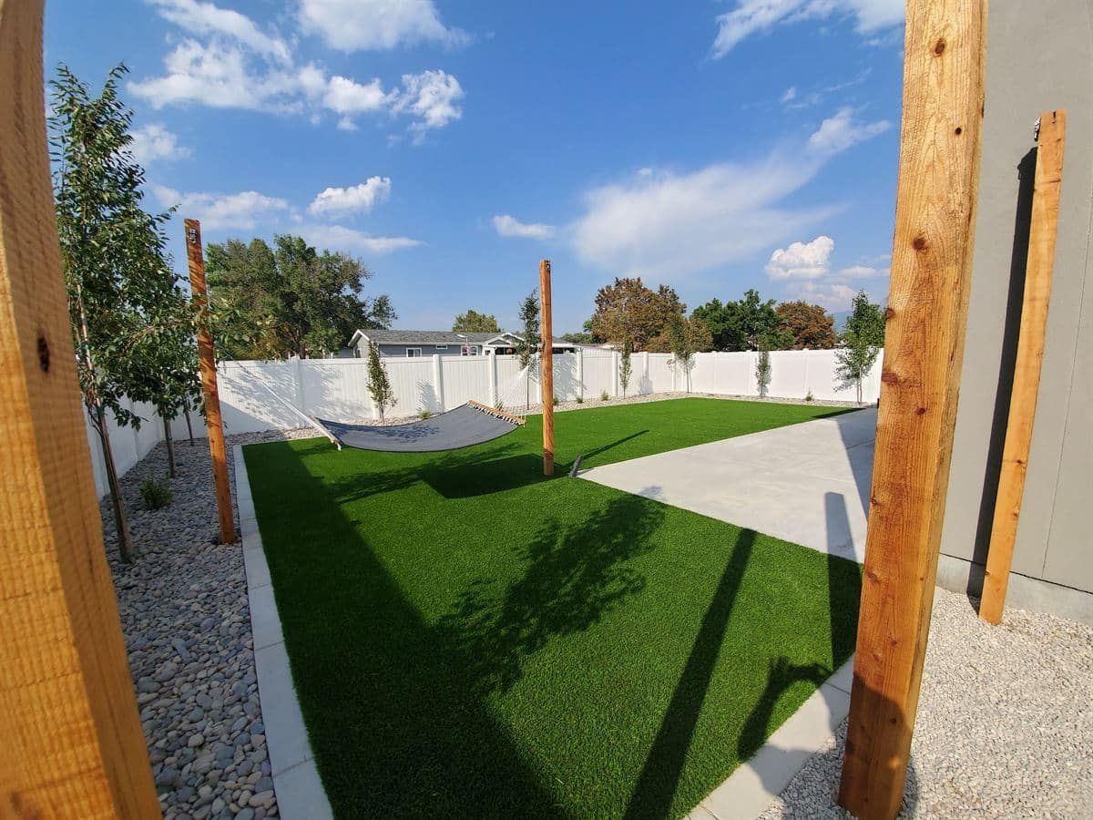 Designed backyard with artificial turf, hammock posts, concrete paving, trees, and white privacy fencing.