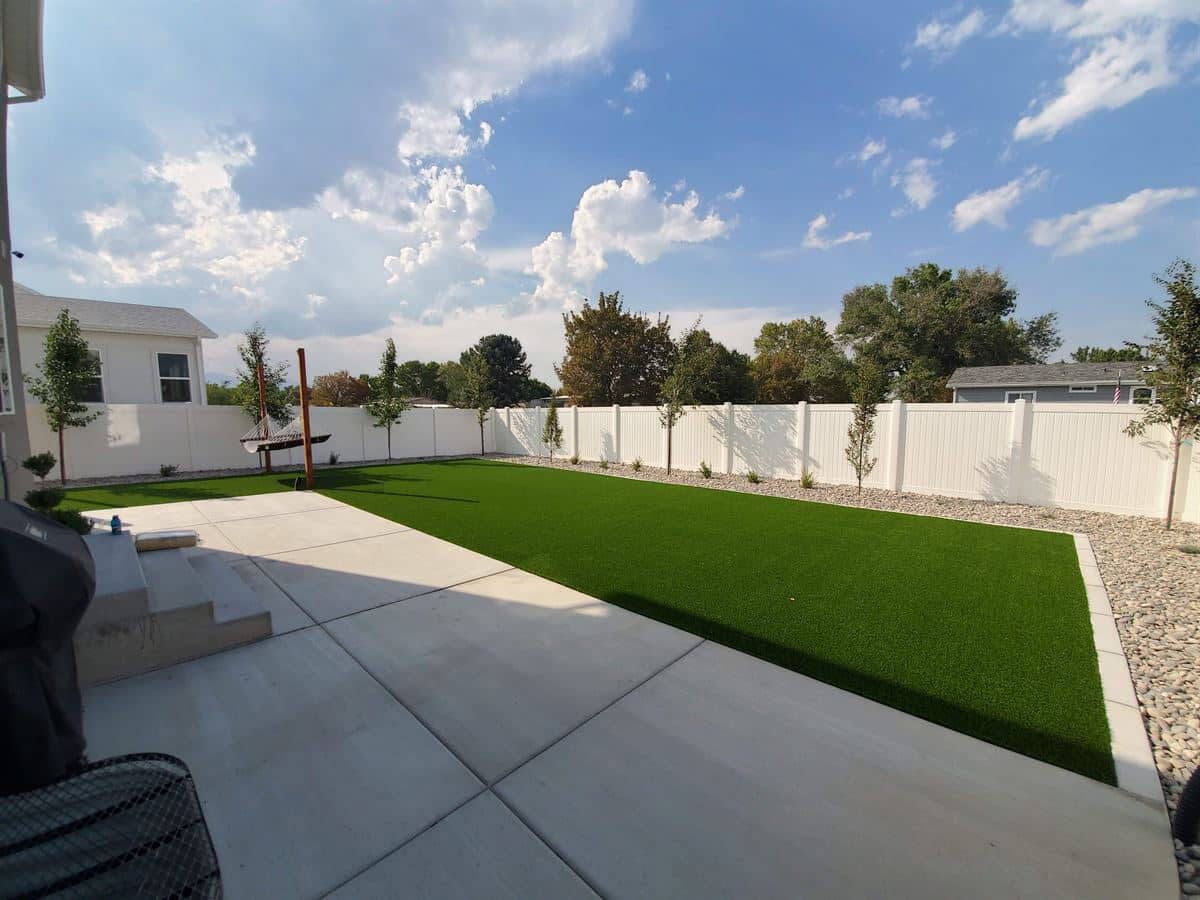 Backyard patio with broad concrete paving, artificial turf, white privacy fence, and young trees.