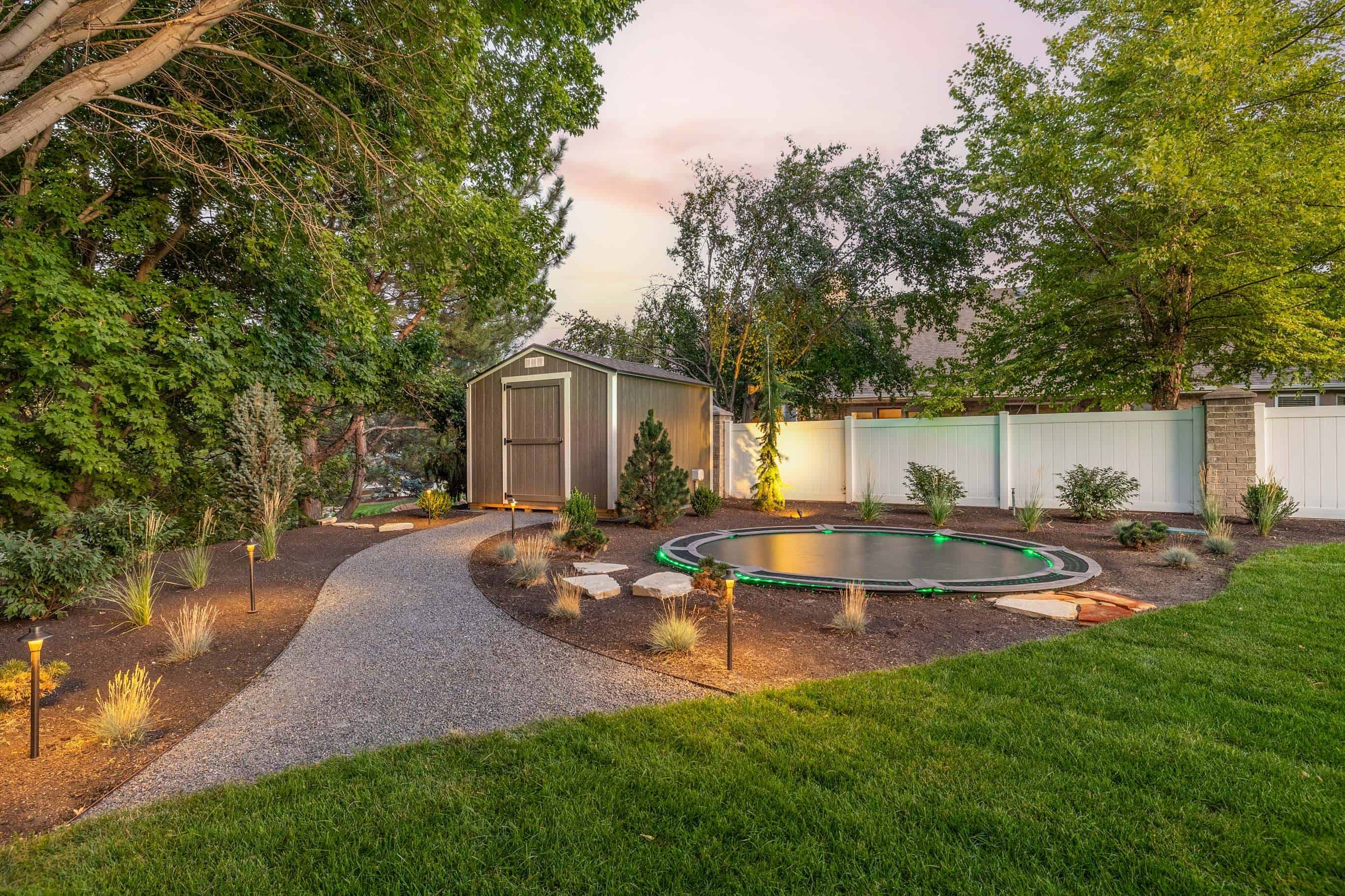 Gravel garden path beside a storage shed, trampoline area, and illuminated planting beds.