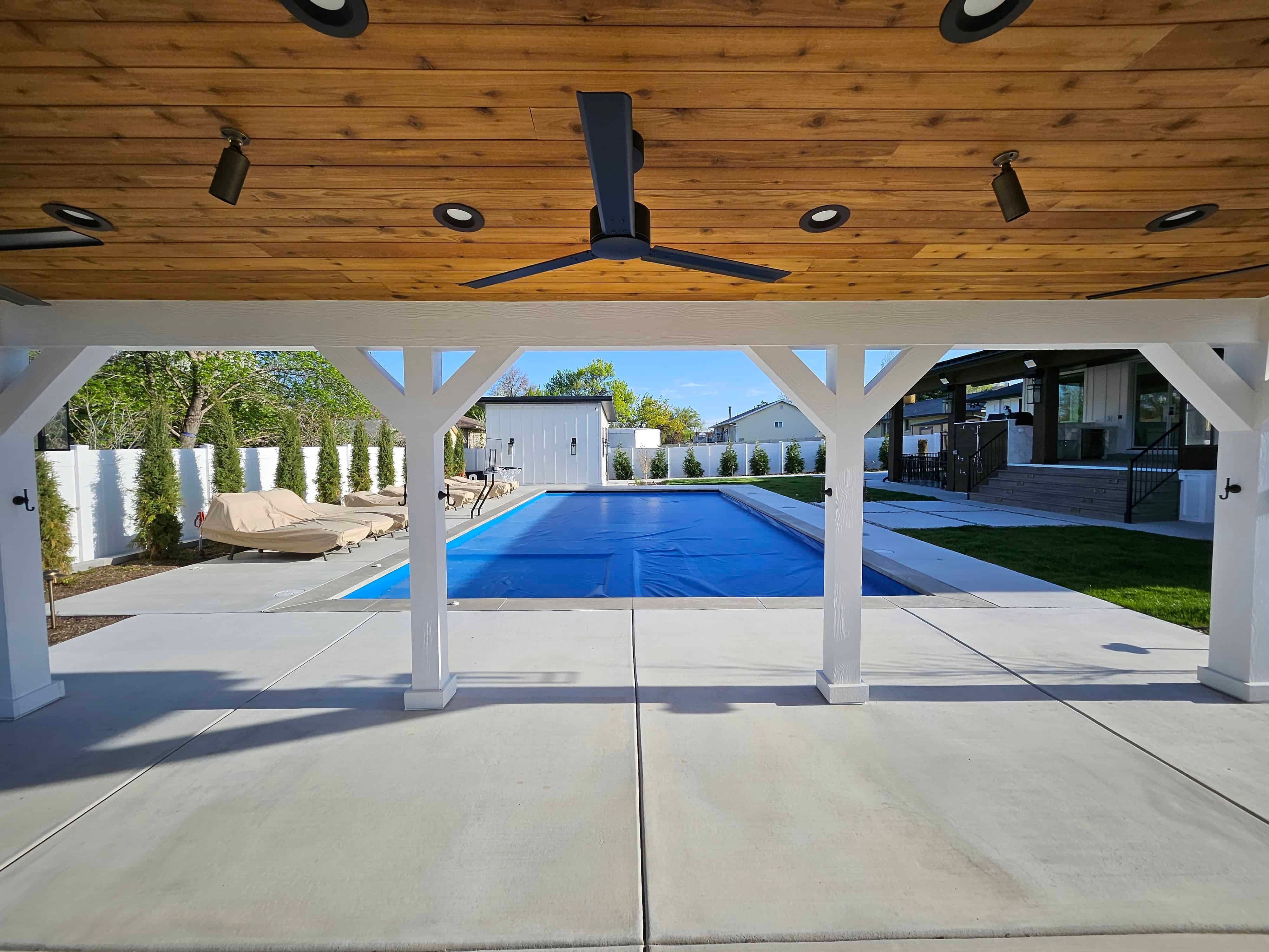 View from under a backyard shade structure looking out to a swimming pool with a concrete deck in South Jordan.
